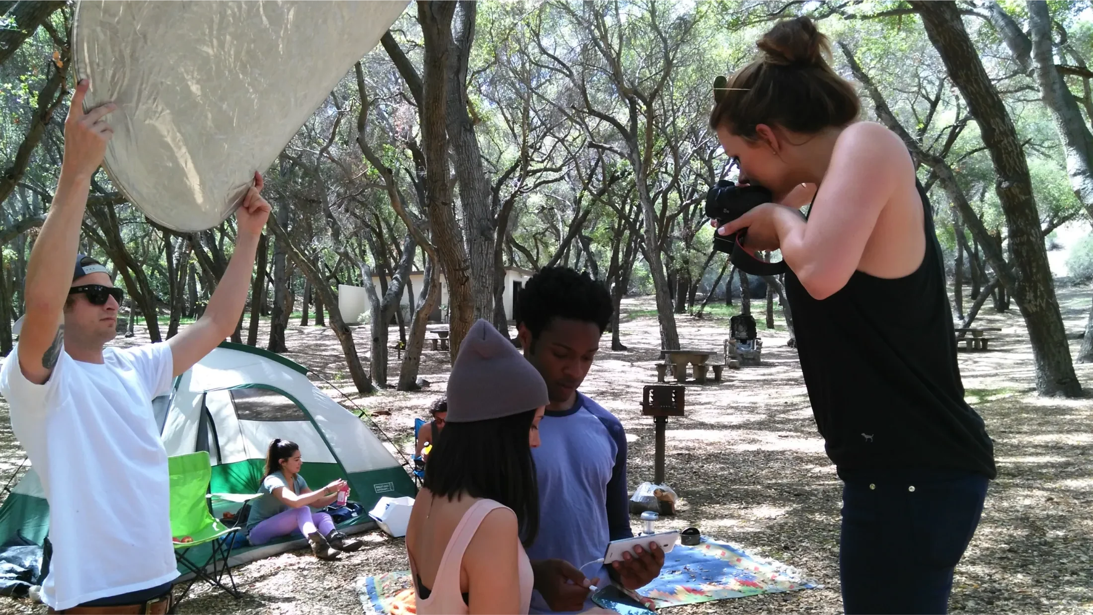 Behind the scenes of the Huawei photoshoot, a photographer is taking a picture of two young adults at a campsite while an assistant holds a diffuser behind them