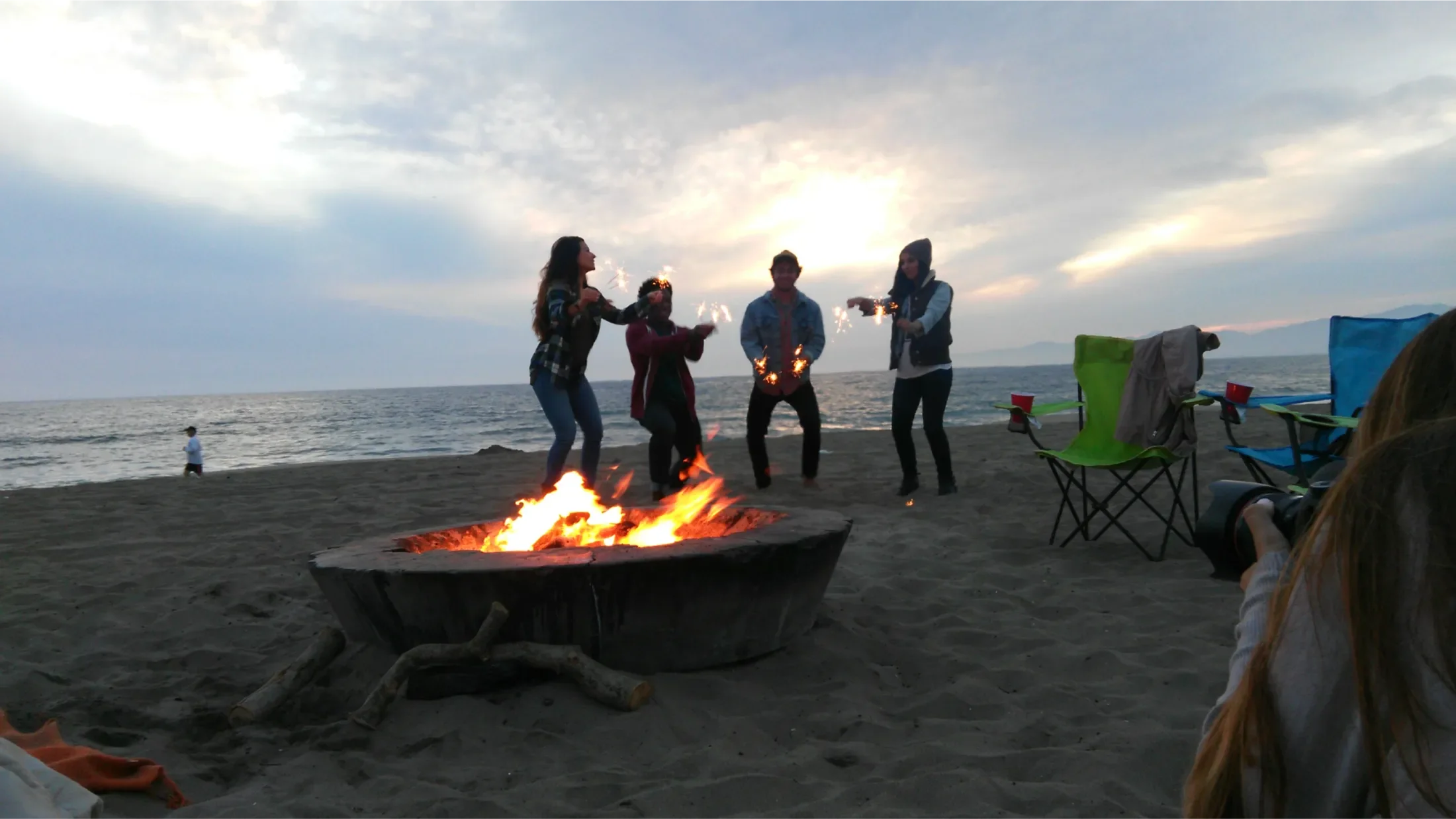 Behind the shoulder of a photographer taking a picture of four young adults on a beach playing with sparklers