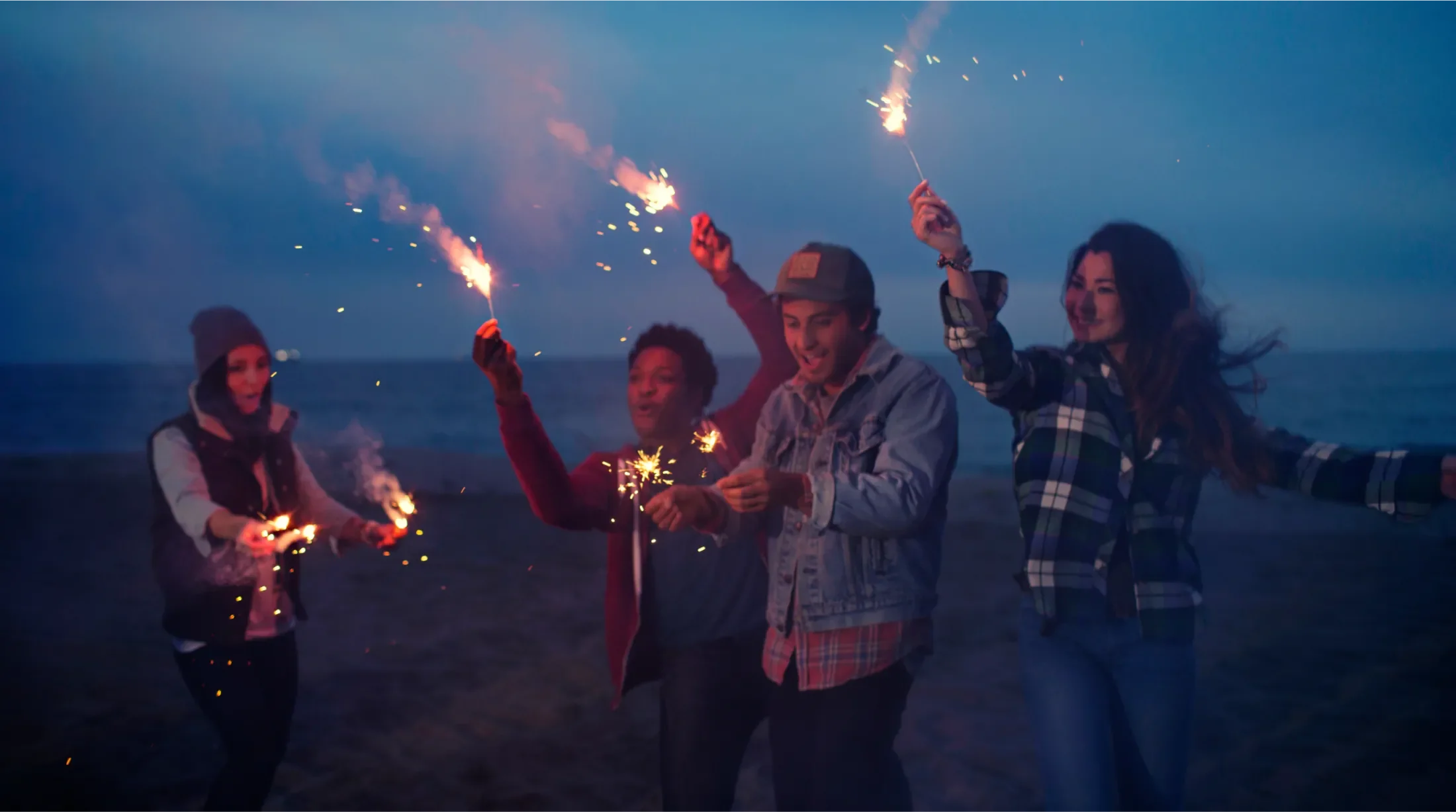 A group of four young adults on a beach at night holding sparklers