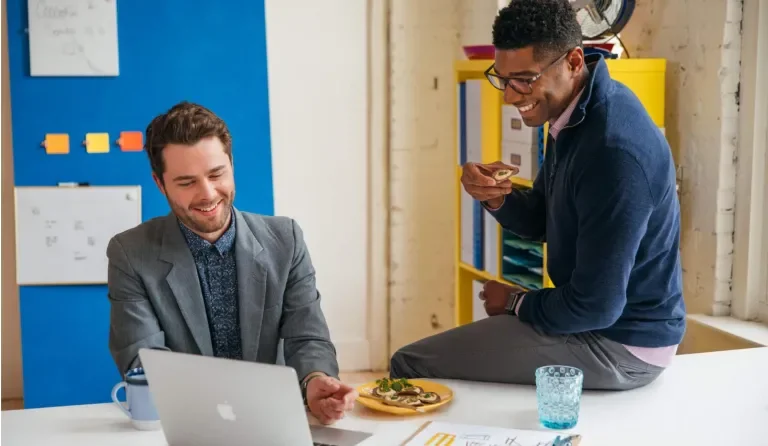 Two smiling men look at a laptop together while one of them enjoys crackers with Laughing Cow cheese on top