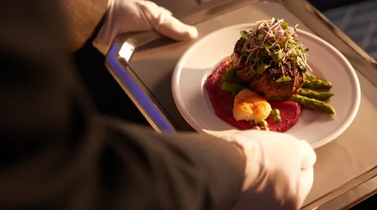 White-glove service, flight attendant serving a filet with asparagus and beet puree topped with microgreens and scallop.