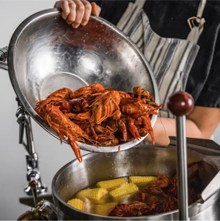 Man pouring a big bowl of lobsters into a large pot full of corn and other seafood