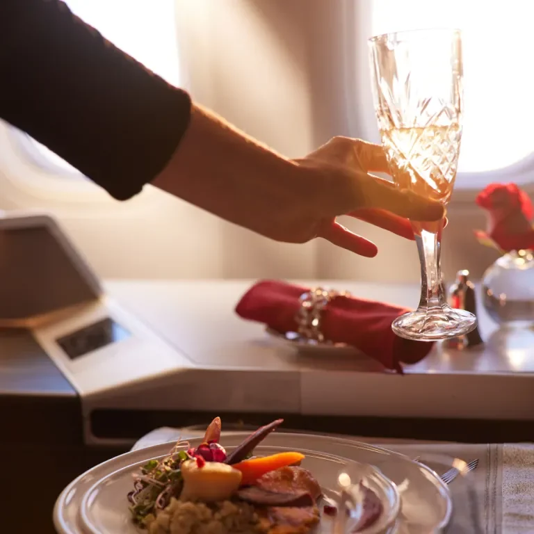 Flight attendant serves champagne in a flute glass in the private suites of a Boeing 777, sunset breaks through aircraft window.